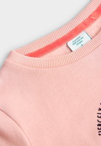 Sudadera rosa claro con cuello y puños acanalados. Presenta una textura suave y una etiqueta con texto azul en el interior del cuello.