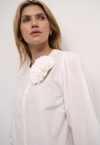 Hvid skjorte med knapper og puffede ærmer, prydet med en tekstureret blomsterdetalje. Stoffet fremstår glat og let, med et klassisk design.