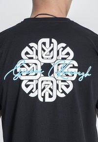 Camiseta negra con un gran diseño gráfico blanco intrincado en la parte posterior, acentuado por texto azul estilizado. Tela suave y texturada.