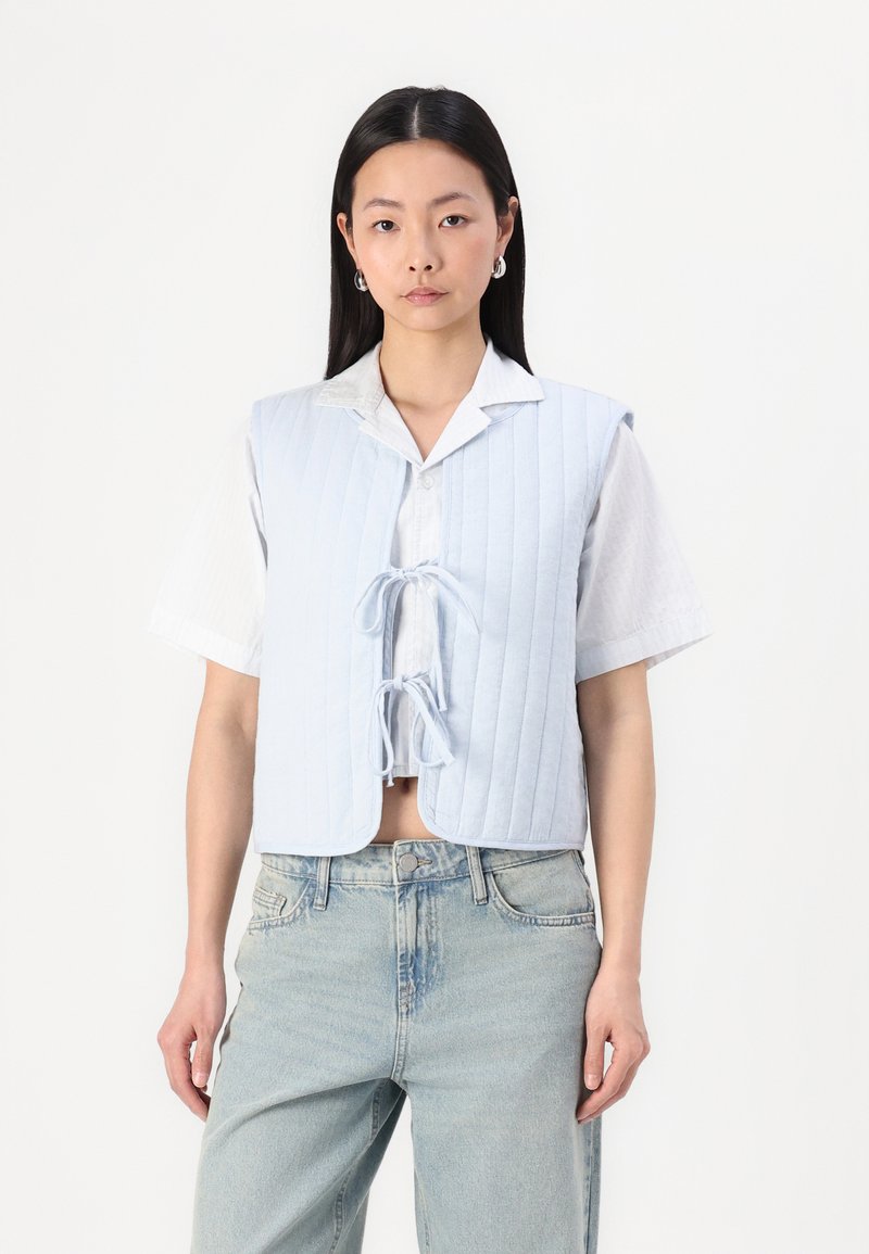 Lyseblå quiltet vest med knytepreg i front, rundede kanter og korte ermer, parret med en hvit skjorte med krage og denimjeans.