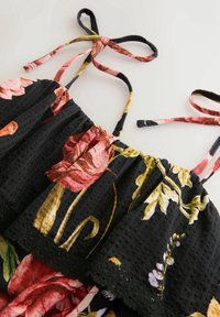 Robe en tissu noir avec un grand imprimé floral rose et jaune, bretelles à nouer, et bord supérieur élastiqué froncé avec détail en dentelle.