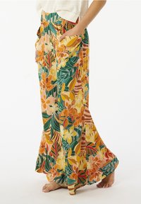 Pantalons larges floraux en orange, jaune et vert avec une texture douce. Présente des poches latérales et une coupe décontractée, mettant en avant un motif tropical.