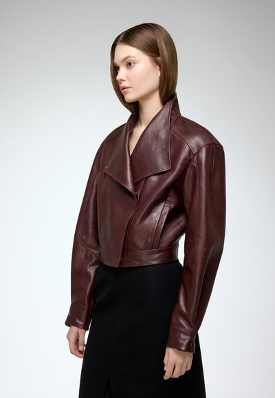 Burgundy leather cropped jacket with wide lapels and asymmetrical zipper. Smooth texture and tailored fit, paired with a black skirt.