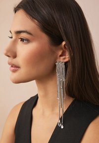 Silver chandelier earrings with long, cascading chains, adorned with clear crystals and two teardrop pendants, worn by a model.