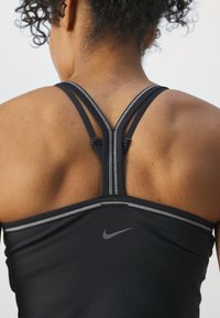 Nainen, jolla on kihara hius ja päällä musta Nike-urheilutoppi, jossa on yksityiskohtaiset ristikkäiset olkahihnat ja näkyvä ihon rakenne harteilla.