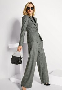 Vrouw in een grijs gestreept pak, zwarte hoge hakken, oversized zonnebril, en met een zwarte handtas van gepatenteerd leer met kettingband.