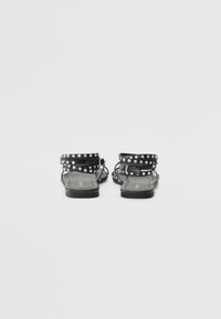 Sandales plates noires avec des sangles cloutées, dotées d'ac accents de boucle. Fabriquées en matériau synthétique avec une texture lisse et une semelle basse.