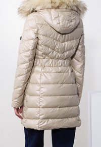Béžový prošívaný zimní kabát s kapucí lemovanou světle hnědým umělým kožešinou, s elastickým pasem, nošený osobou stojící na bílém pozadí.