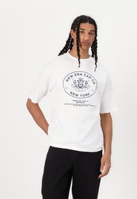Hvid bomulds-T-shirt med et marineblåt grafisk tryk af "New Era Cap Co" og et løveemblem, med korte ærmer og en afslappet pasform.