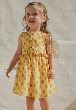 Naeratav noor tüdruk pikkade juustega, kes kannab kollast lillemustriga varrukateta kleiti, seisab valge tekstureeritud seina vastu.