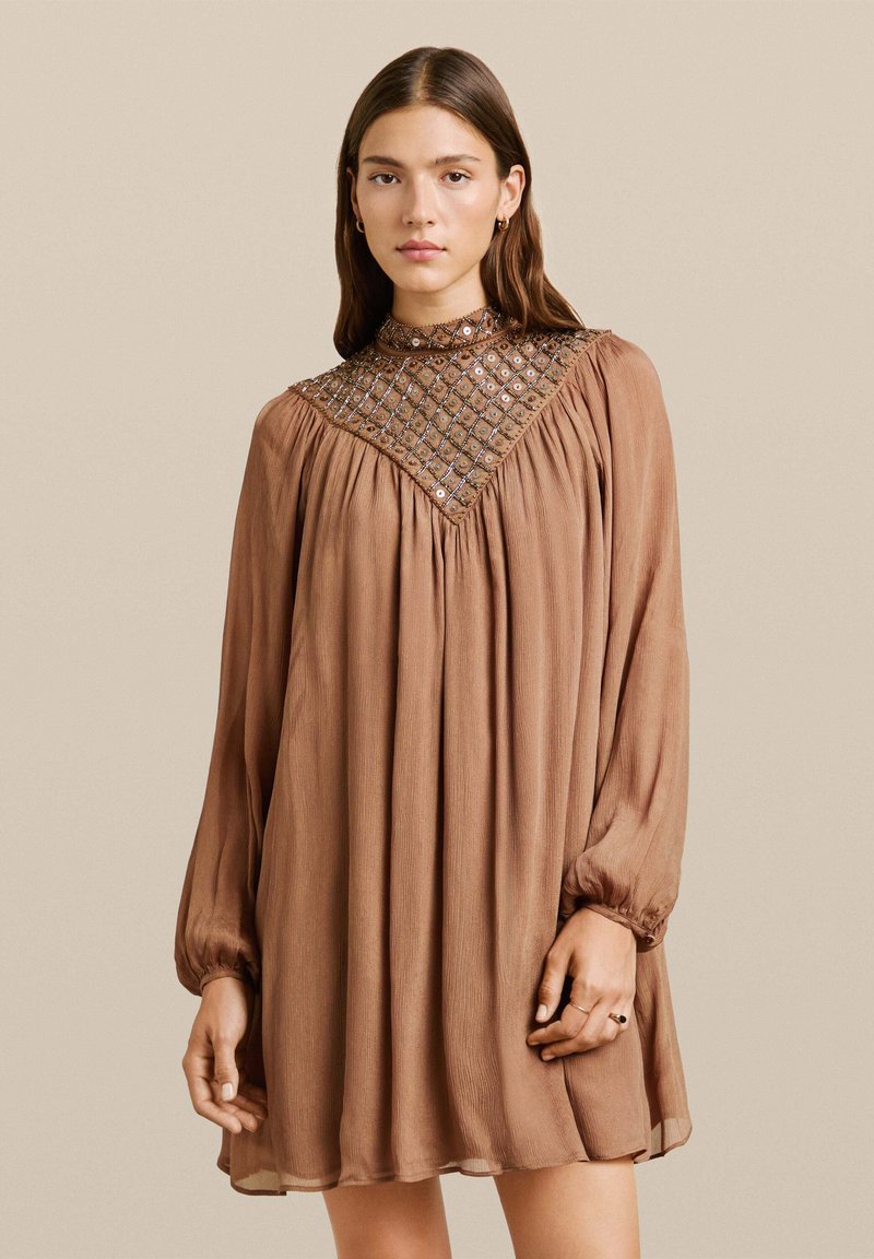 Vestido marrón de manga larga con diseño plisado y acentos de malla con cuentas. Cuello alto y corte holgado, hecho de una tela ligera y texturizada.