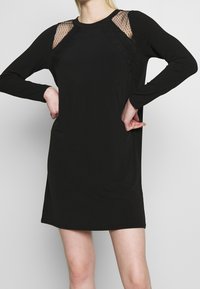 Musta pitkähihaista mekkoa, jossa on läpinäkyvät pitsikoristeet olkapäillä ja pyöreä kaula-aukko. Sujuva kangas, rento malli ja keskimmäisen reiden pituus.