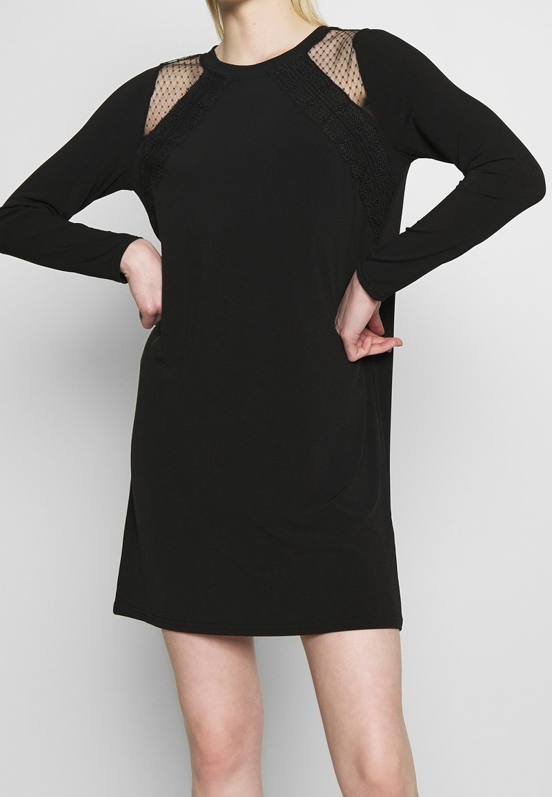 Musta pitkähihaista mekkoa, jossa on läpinäkyvät pitsikoristeet olkapäillä ja pyöreä kaula-aukko. Sujuva kangas, rento malli ja keskimmäisen reiden pituus.