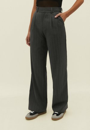 Vrouw draagt hooggetailleerde, donkergrijze wijduitlopende pantalon met krijtstrepen, riem en zwart-grijze sneakers, staand met één hand in de zak.