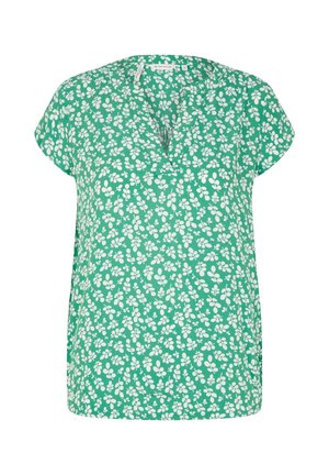 Kvinde iført mørkeblå denimjakke, grøn blomsterbluse, vidde blå jeans og hvide sneakers står mod en hvid baggrund.