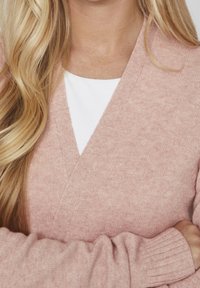 Ljusrosa, stickad cardigan med V-ringning och ribbade manschetter, lager på lager över en vit topp. Mjuk textur, avslappnad passform, subtila mönster.