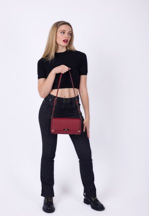 Rode leren schoudertas met een platte ontwerp, metalen accenten en een schouderband. Model in een zwart crop top en donkere spijkerbroek, staand.