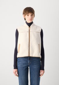 Off-white fleece vest with brown leather accents, zip closure, standing collar, and side pockets. Worn over a navy long-sleeve top.