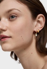 Goldfarbene Ohrringe mit einem floralen Anhänger und glattem, strukturierter Ring, mit einem blauen Akzent. Nahaufnahme von Ohrschmuck an einem Modell.