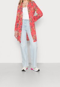 Rød blomsterkimono med lange ærmer, parret med lyseblå højtaljede jeans og farverige sneakers. Baggrunden er helt hvid.