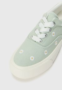 Light green canvas sneaker with white laces, featuring white embroidered flowers with yellow centers, rubber sole, and textured toe cap.