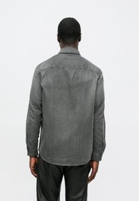 Bărbat îmbrăcat cu o cămașă gri cu mânecă lungă și pantaloni negri, stând cu spatele, pe un fundal alb simplu.