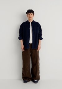 Jeune homme debout contre un mur blanc, portant une chemise bleu marine, un t-shirt blanc, un pantalon large marron et des baskets foncées.