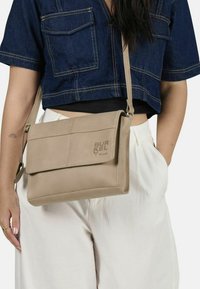 Borsa a tracolla beige con chiusura a pattina, texture liscia, dettaglio del logo e tracolla regolabile. Indossata con una maglietta di jeans e pantaloni chiari.