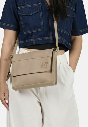 Beige Umhängetasche mit Klappenverschluss, glatter Textur, Logo-Detail und verstellbarem Trageriemen. Getragen mit einem Denim-Oberteil und hellen Hosen.