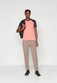 Top desportivo de manga curta rosa e preto, calças bege, segurando uma mochila preta, em pé sobre um fundo simples.