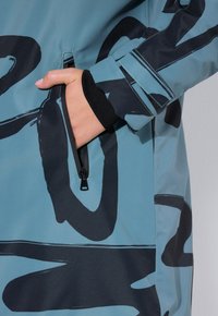 Giacca impermeabile blu con audaci motivi astratti neri. Presenta una tasca con zip e polsini a coste per una maggiore presa. Texture liscia.