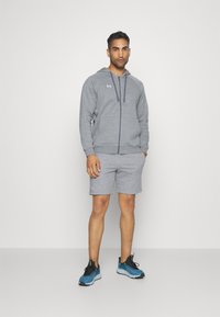 Grå hettegenser med snor og glidelås, kombinert med grå shorts. Modellen har på seg blå joggesko med svarte accenter. Minimalistisk design.
