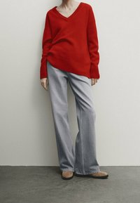 Pull en maille rouge avec un col en V et un ourlet côtelé, associé à un jean large gris clair et des ballerines beiges. Textures lisses, coupe décontractée.