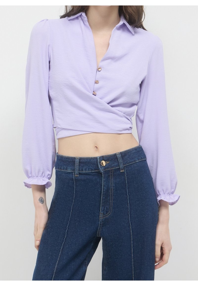 Femme portant un chemisier court style cache-cœur lavande avec manches bouffantes et un jean taille haute bleu foncé avec coutures devant.