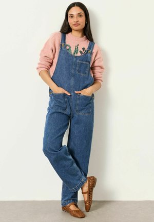 Woman with long dark hair wearing pink sweater, blue denim overalls, and brown woven flats, standing against a white wall with hands in pockets.