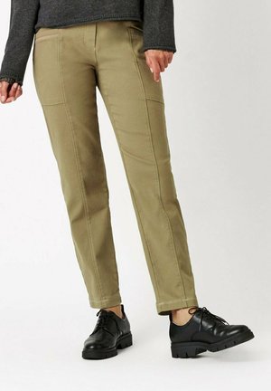 Khakihosen mit Seitennähten, vorderen Taschen und dezenten Nähten. Kombiniert mit schwarzen Lederschuhen mit dicker Sohle und Schnürsenkeln.