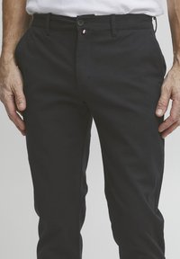 Melnas kokvilnas bikses ar gludu tekstūru, priekšējo pogu aizdari un nelielu trīs krāsu akcentu tuvumā jostasvietai. Pieguļoša piegriezuma dizains.