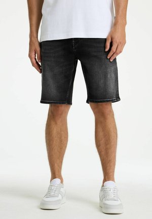 Man wearing black denim shorts, white sneakers, and a white t-shirt, standing against a plain white background.