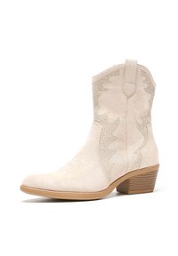 Botas de cowboy de ante beige con costuras decorativas y patrones perforados. Presentan una punta afilada y un tacón apilado.