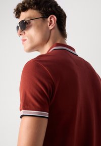Crvena polo majica s rebrastom teksturom, s bijelim i crnim prugastim detaljima na ovratniku i rukavima. Model nosi sunčane naočale.