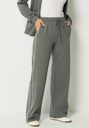 Persona che indossa pantaloni della tuta grigi a gamba larga con strisce laterali bianche, top bianco, giacca grigia, mani in tasca e scarpe bianche.
