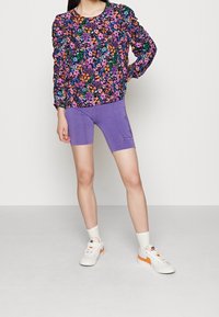 Personne portant un chemisier à fleurs coloré à manches longues, un short de cycliste violet, des chaussettes blanches et des baskets blanches avec des accents orange, debout de face.