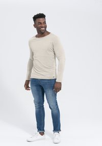 Helle beige Langarmbluse mit rundem Ausschnitt, kombiniert mit blauen Slim-Fit-Jeans und weißen Sneakers. Glatte Textur, minimalistisches Design.