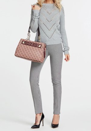 Femme portant un pull gris à motifs et un pantalon, tenant un sac à main rose de la marque Guess, portant des escarpins noirs avec un tatouage floral visible à la cheville.