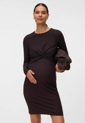Femme enceinte dans une robe sombre ajustée avec un détail noué, tenant une veste pliée dans une main et posant l'autre sur son ventre.