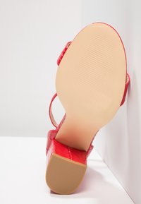 Sandale rouge à talon haut avec semelle texturée et talon bloc, inclinée vers le haut contre une surface blanche en coin.
