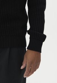 Högra armen på en person som bär en svart texturerad tröja med uppkavlade ärmar och svarta byxor mot en vit bakgrund.