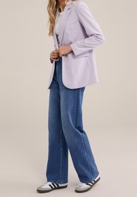 Kvinna som knäpper en ljuslila blazer över en vit topp, klädd i högmidjade blå vida jeans och vita sneakers med svarta ränder.