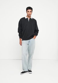 Ung mann står med den ene hånden i lommen, iført svart langermet skjorte med krage, lyseblå jeans og svarte joggesko mot hvit bakgrunn.