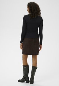 Top negru cu mânecă lungă, fustă din suede maro, cizme negre până la genunchi cu catarame; păr creț, văzut din spate pe un fundal deschis.